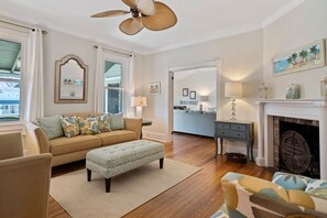 A living room with a beige sofa, patterned ottoman, wooden flooring, and a fireplace.