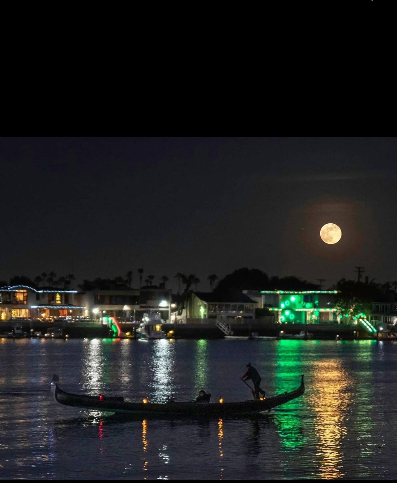 Escena nocturna con luna llena, una góndola en el agua y edificios iluminados al fondo.