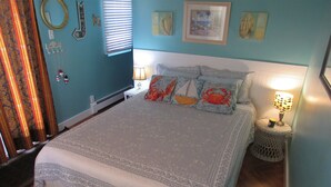 A bedroom with a bed, a nightstand with a lamp, a window with blinds, and a wall with framed pictures.