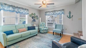 Una sala de estar con un sofá azul claro, dos sillones, un tapete estampado y decoración náutica que incluye un ventilador de techo y cortinas de ventana.