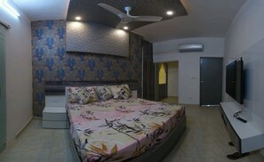 Relax in this serene bedroom featuring a large bed with a floral bedspread, ceiling fan, air conditioning, and a wall-mounted TV.