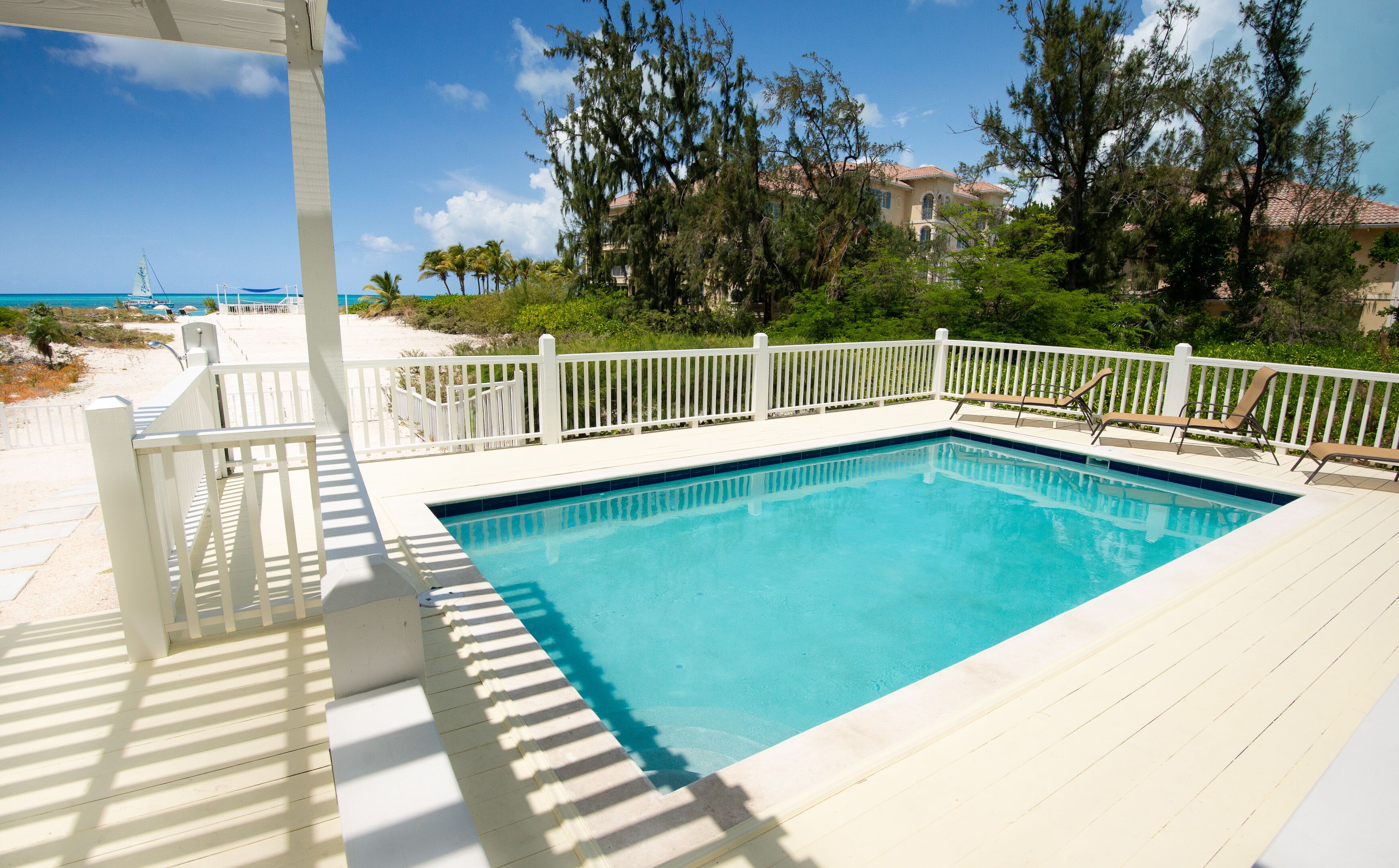A clear blue pool surrounded by a white deck with a white fence, overlooking a beach and ocean.