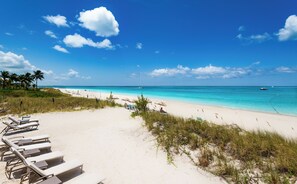 A beach with white sand, clear turquoise water, and a few boats. There are lounge chairs and umbrellas along the shore.