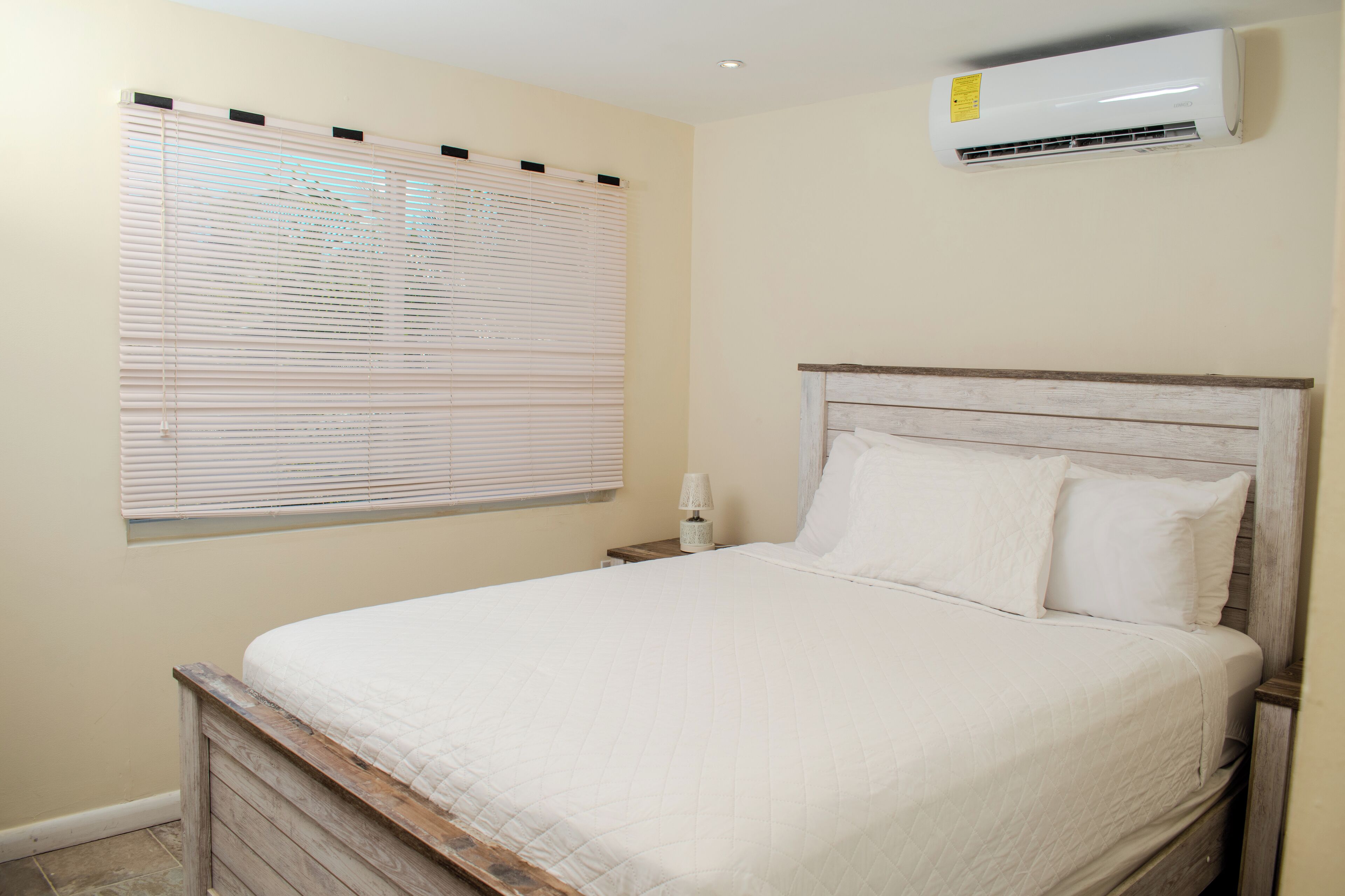 A bedroom with a wooden bed frame, white bedding, a bedside lamp, and an air conditioning unit on the wall.