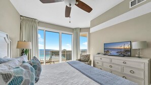 Habitación de hotel con una cama grande, ventilador de techo, televisor, cómoda y balcón con vista al océano.