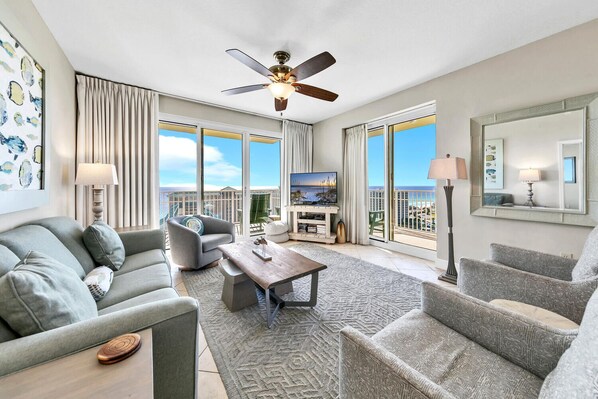 A spacious living room with a ceiling fan, a flat-screen TV, and a balcony with a view of the ocean.
