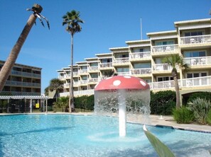 Ein Swimmingpool mit Wasserspiel in der Mitte, umgeben von Palmen und einem Gebäude mit Balkonen.
