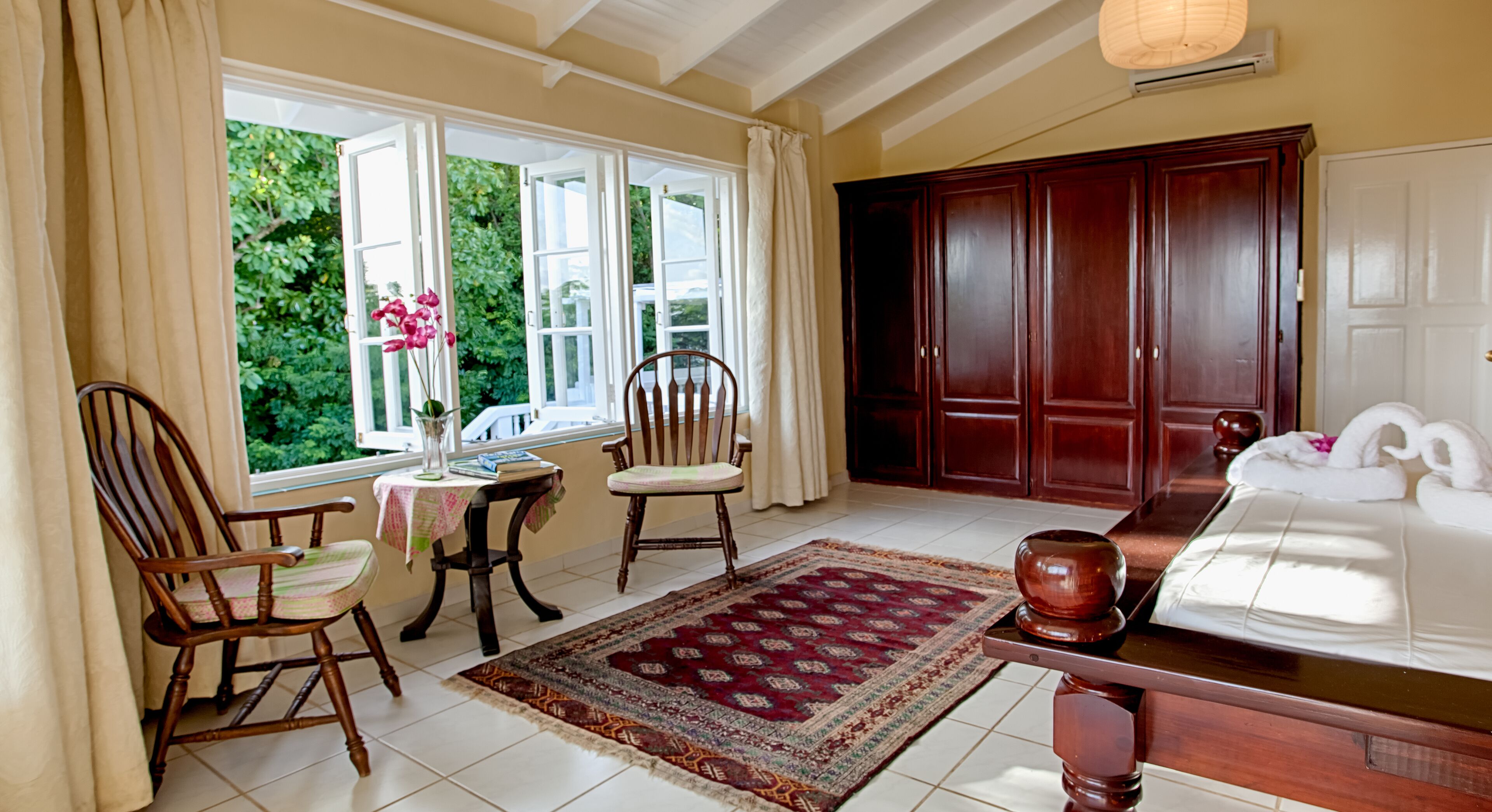 Une pièce comprenant une grande armoire en bois, un lit avec du linge de maison blanc, un fauteuil à bascule en bois, une petite table avec un vase de fleurs et une vue sur la verdure par les fenêtres.