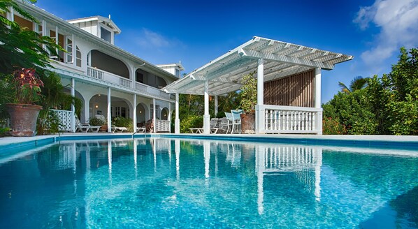 A clear blue pool with lounge chairs and a covered seating area, surrounded by lush greenery and a white building with arches.