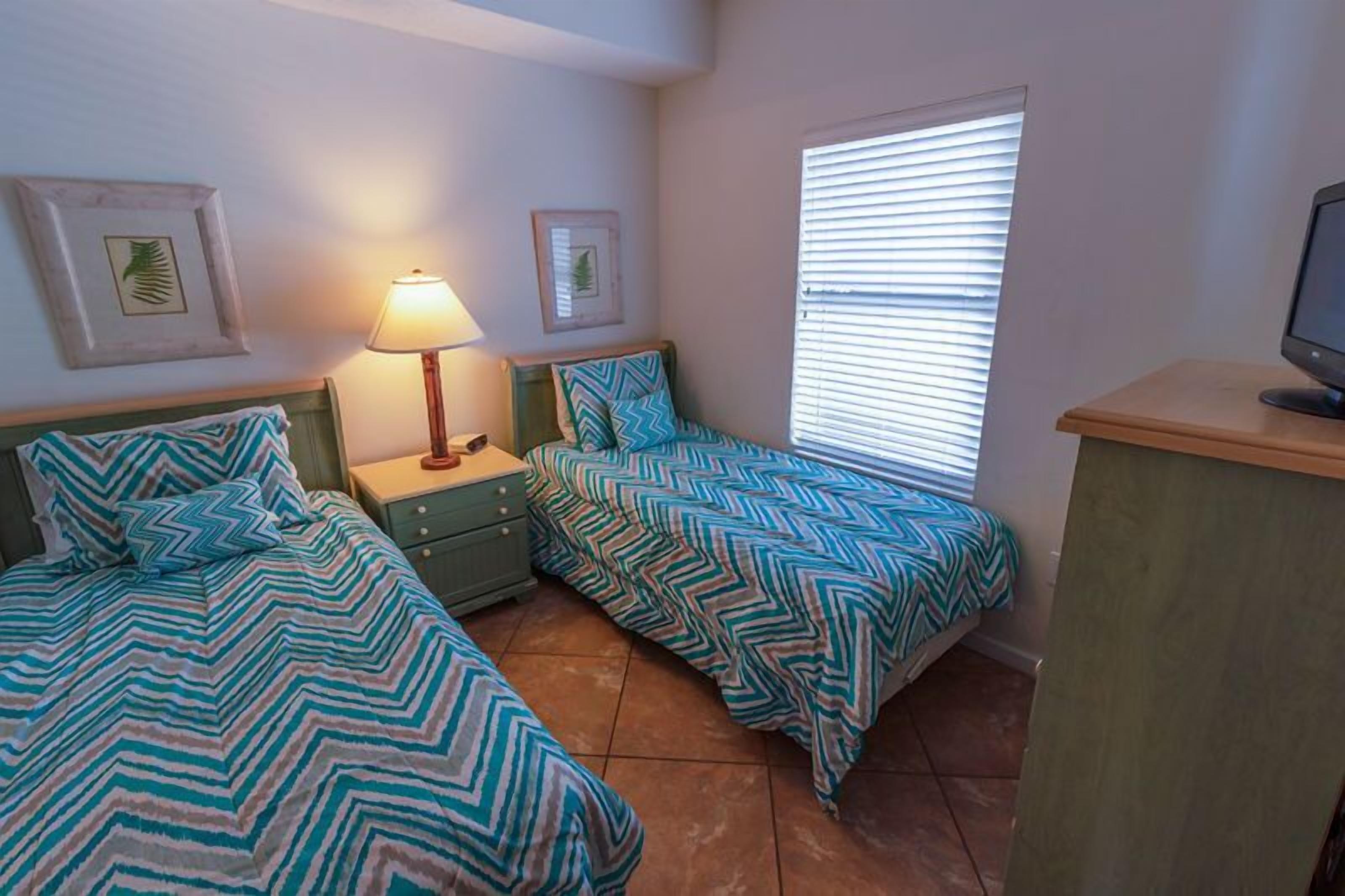 A bedroom with two beds, a nightstand, a lamp, a television, and a window with blinds.