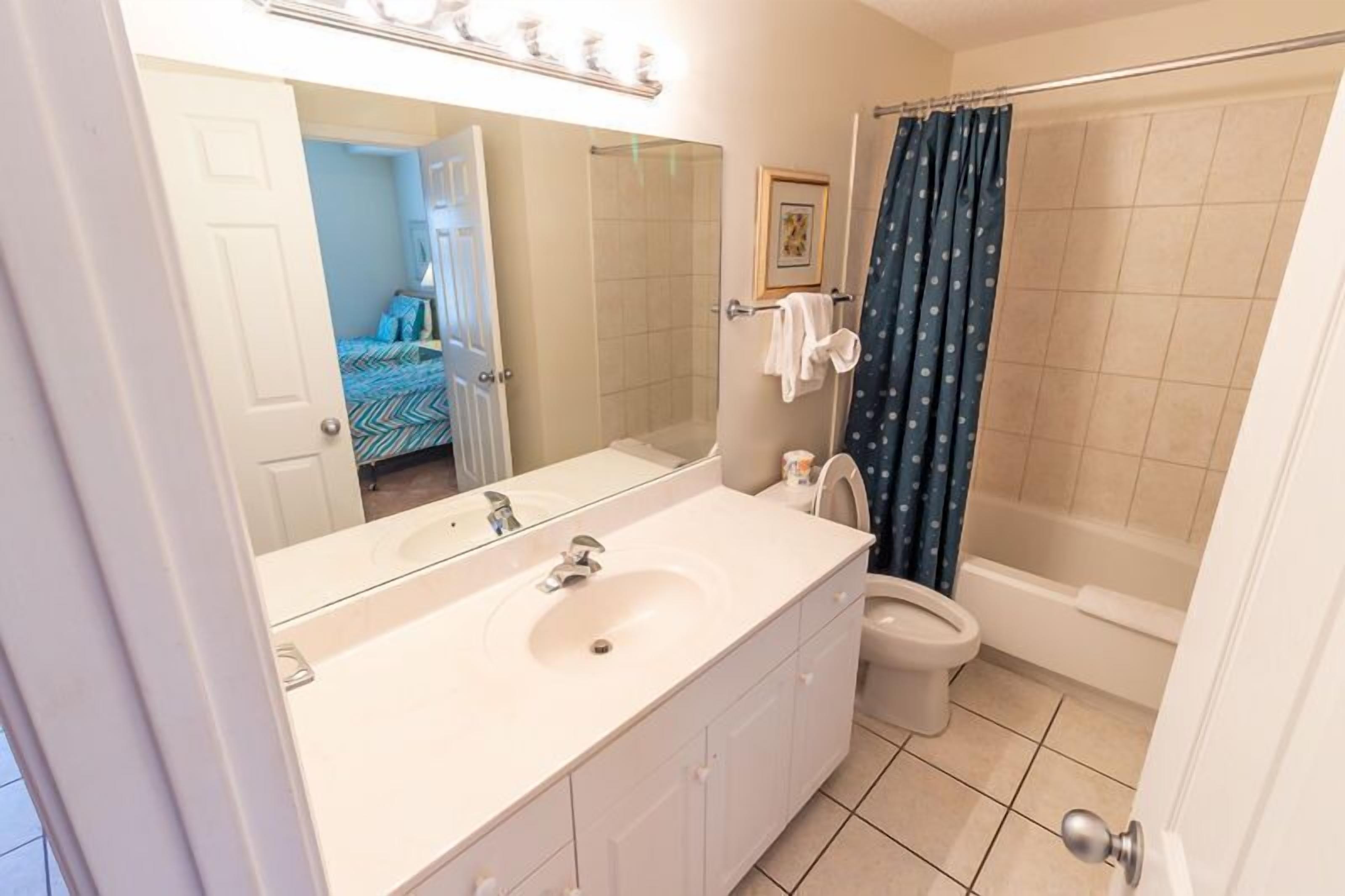 Een badkamer met een wit wastafel, spiegel en toilet. Er is een douchegordijn en een deur die naar een slaapkamer leidt.
