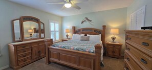 Une chambre à coucher avec un lit en bois, deux tables de chevet, une commode, un ventilateur de plafond et un élément décoratif fixé au mur.
