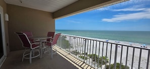 A balcony with a view of the beach, a table, and two chairs.