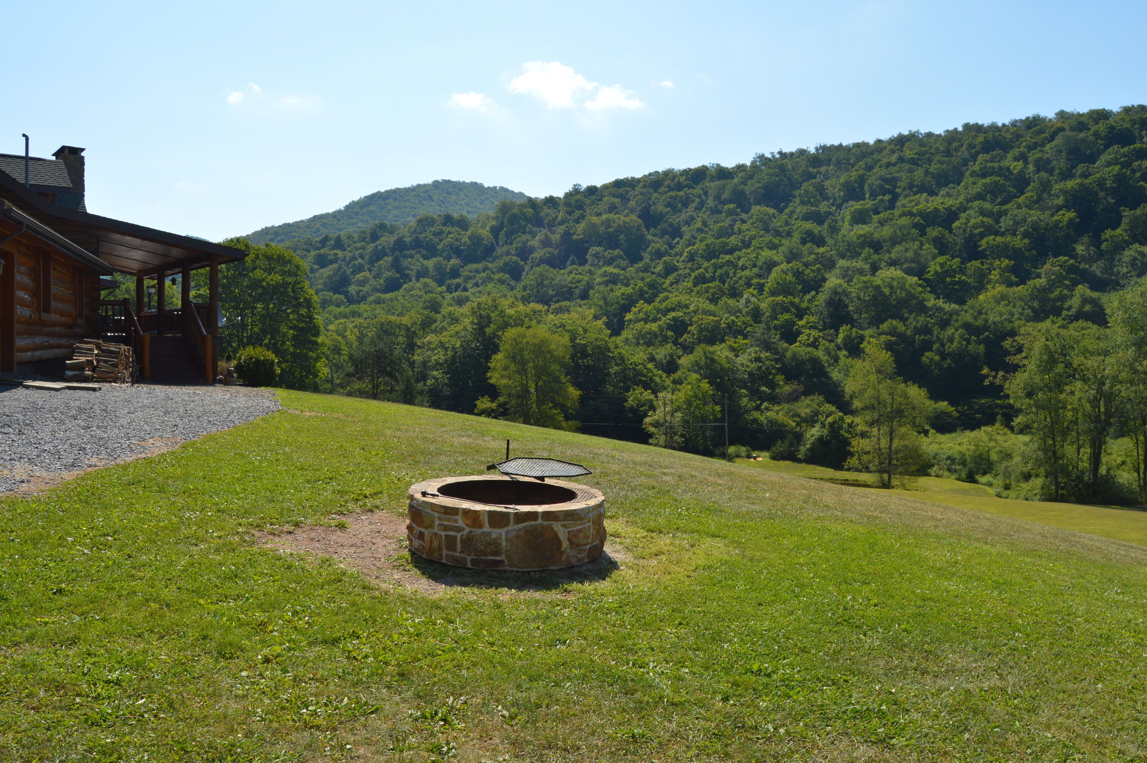 Une cabane rustique avec un foyer en pierre, située dans une zone herbeuse et offrant une vue sur une colline boisée.