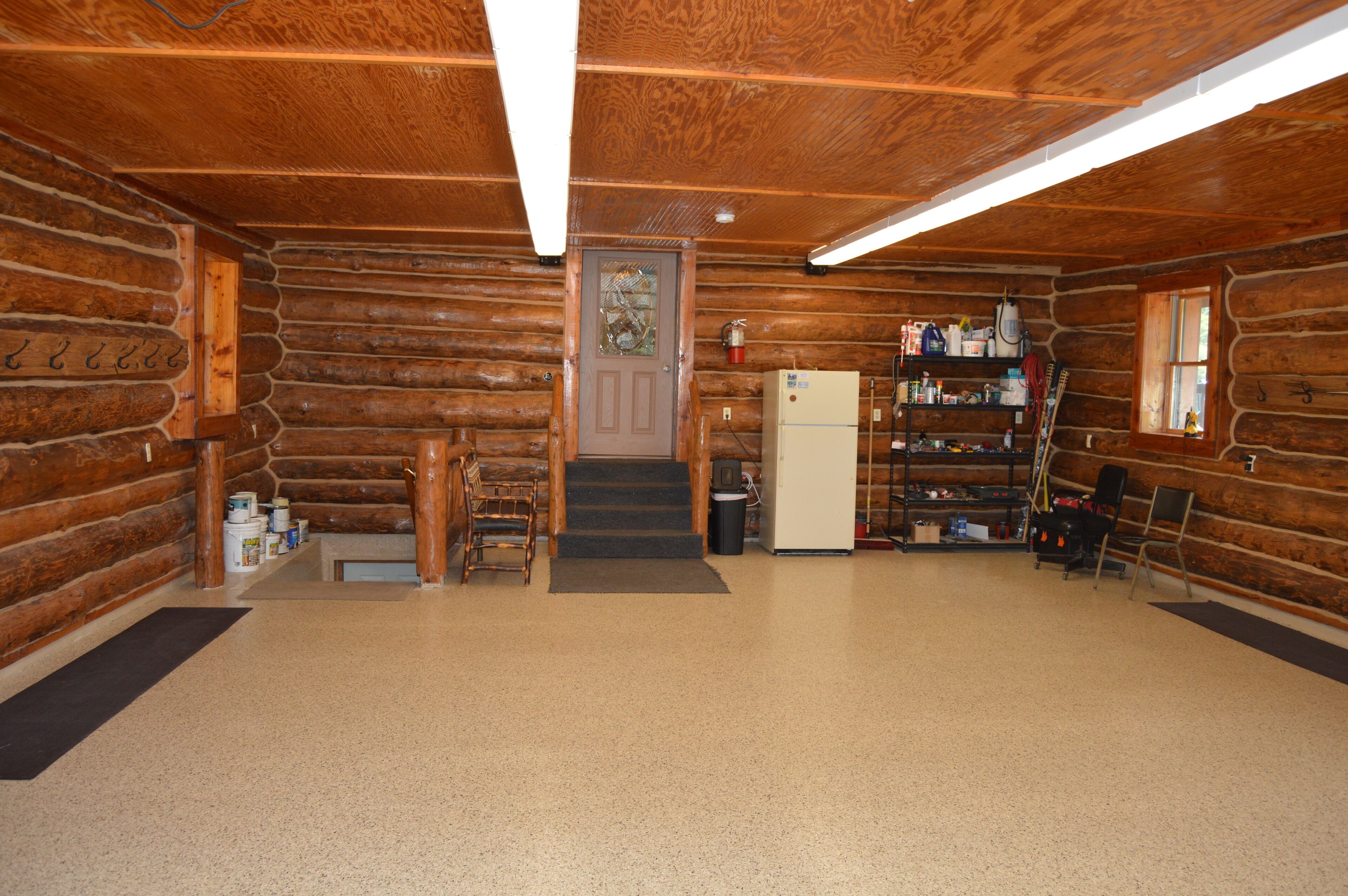 A log cabin interior with a wooden floor, a refrigerator, a staircase, and a shelf with various items.