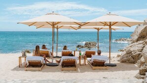 En lounge ved stranden med træliggestole og beige parasoller, med udsigt over havet.