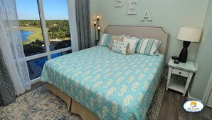 A bedroom with a large window, a bed with patterned bedding, a nightstand with a lamp, and a wall with the word "SEA" spelled out. - True 1 BR with full kitchen, Luau 6532 by Skis and Seas | South Gulf facing (Destin)