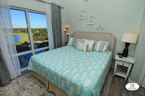A bedroom with a large window, a bed with patterned bedding, a nightstand with a lamp, and a wall with the word "SEA" spelled out. - True 1 BR with full kitchen, Luau 6532 by Skis and Seas | South Gulf facing (Destin)