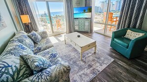 A living room with a sofa, coffee table, and a teal armchair. A television is mounted on the wall, and there is a balcony with a view of buildings and palm trees. - True 1 BR with full kitchen, Luau 6532 by Skis and Seas | South Gulf facing (Destin)