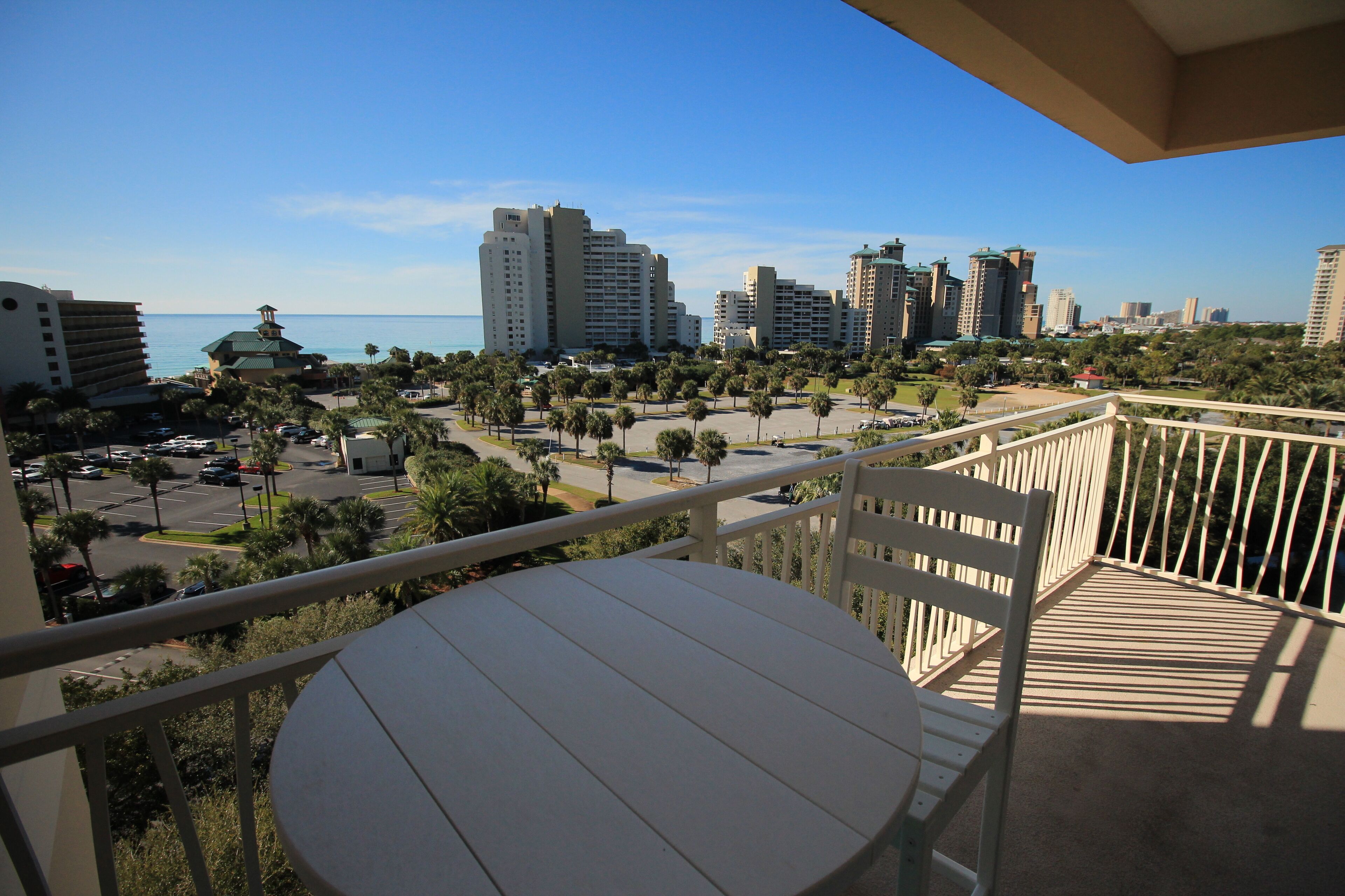 A balcony with a round table and a chair, overlooking a cityscape with tall buildings and a clear blue sky.