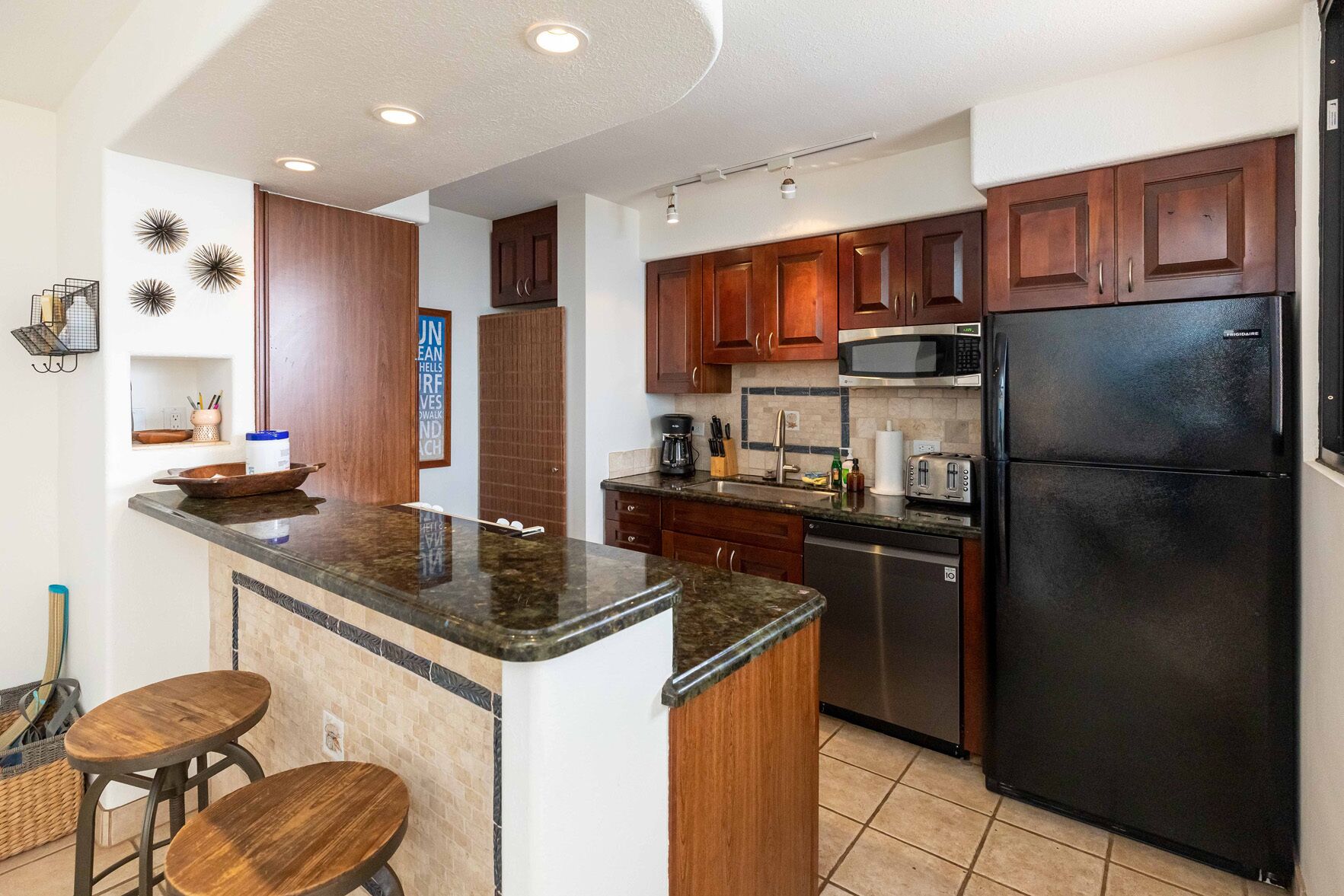A modern kitchen with wooden cabinets, a black refrigerator, a microwave, and a granite countertop.