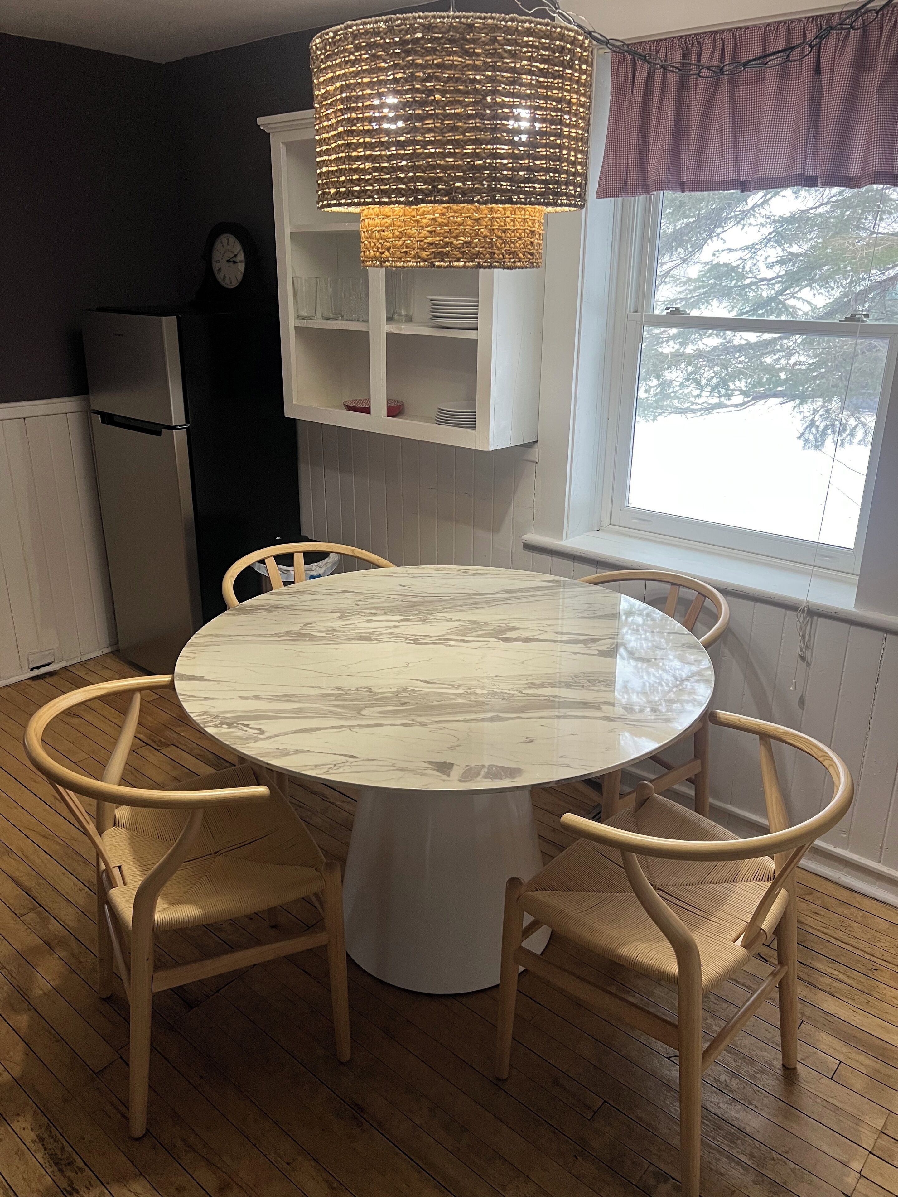 A dining area with a round table and four chairs, a hanging light fixture, a clock, and a window with a view of trees.