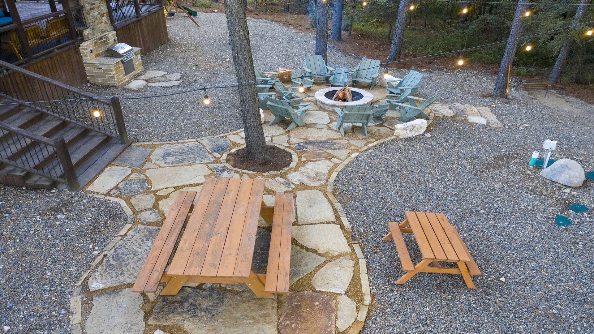 A stone patio with wooden benches and a fire pit surrounded by Adirondack chairs.