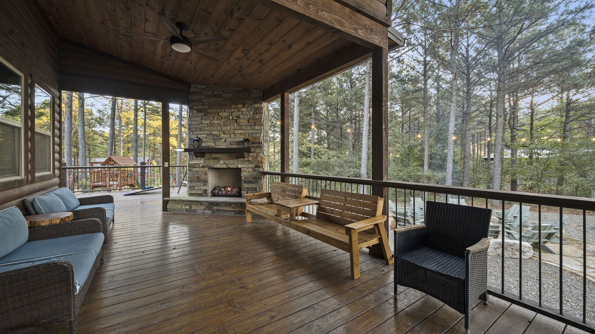 A wooden deck with a stone fireplace, wicker furniture, and a view of a forest.