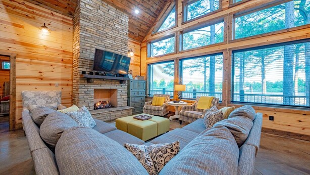 L’intérieur chaleureux d’un chalet avec une cheminée en pierre, des sièges confortables et de grandes fenêtres offrant une vue sur la forêt environnante.