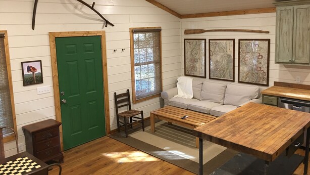 Ein gemütliches Blockhaus-Interieur mit Holzbalkendecke, einer grünen Tür, einem Sofa, einem Esstisch und einem Schachbrett.