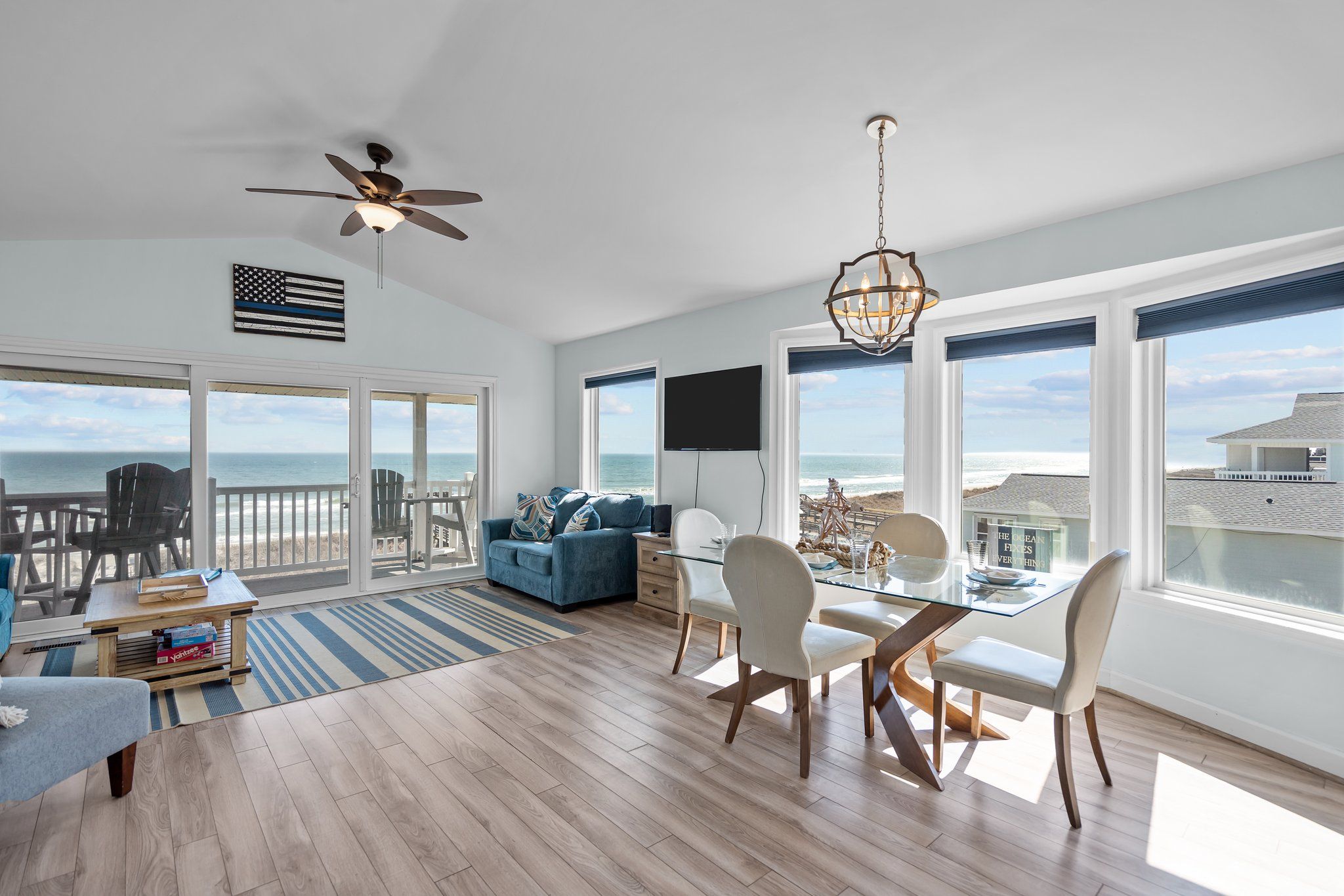 A spacious living area with a balcony overlooking the ocean, a dining table with chairs, a ceiling fan, and a chandelier.