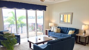 A living room with a blue sofa, a coffee table, and a balcony with a view of greenery and buildings.