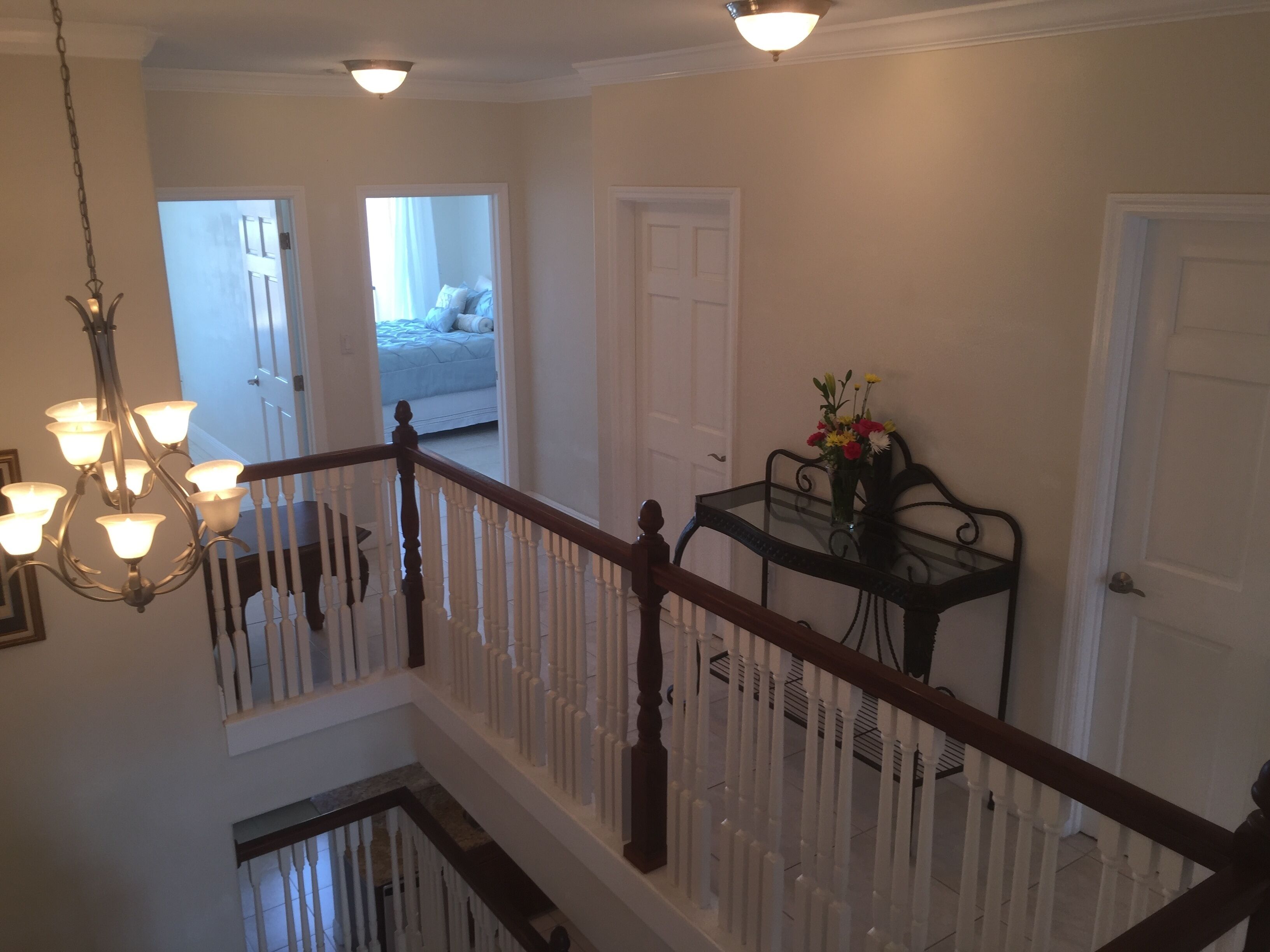 A multi-level interior with a staircase, a chandelier, a dining table with a vase of flowers, and a bed in the background.