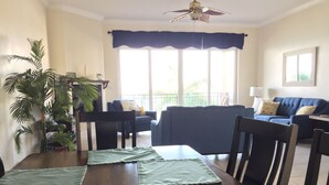 A dining area with a wooden table, green placemats, and dark chairs. A blue sofa and a small table with a lamp are in the background.