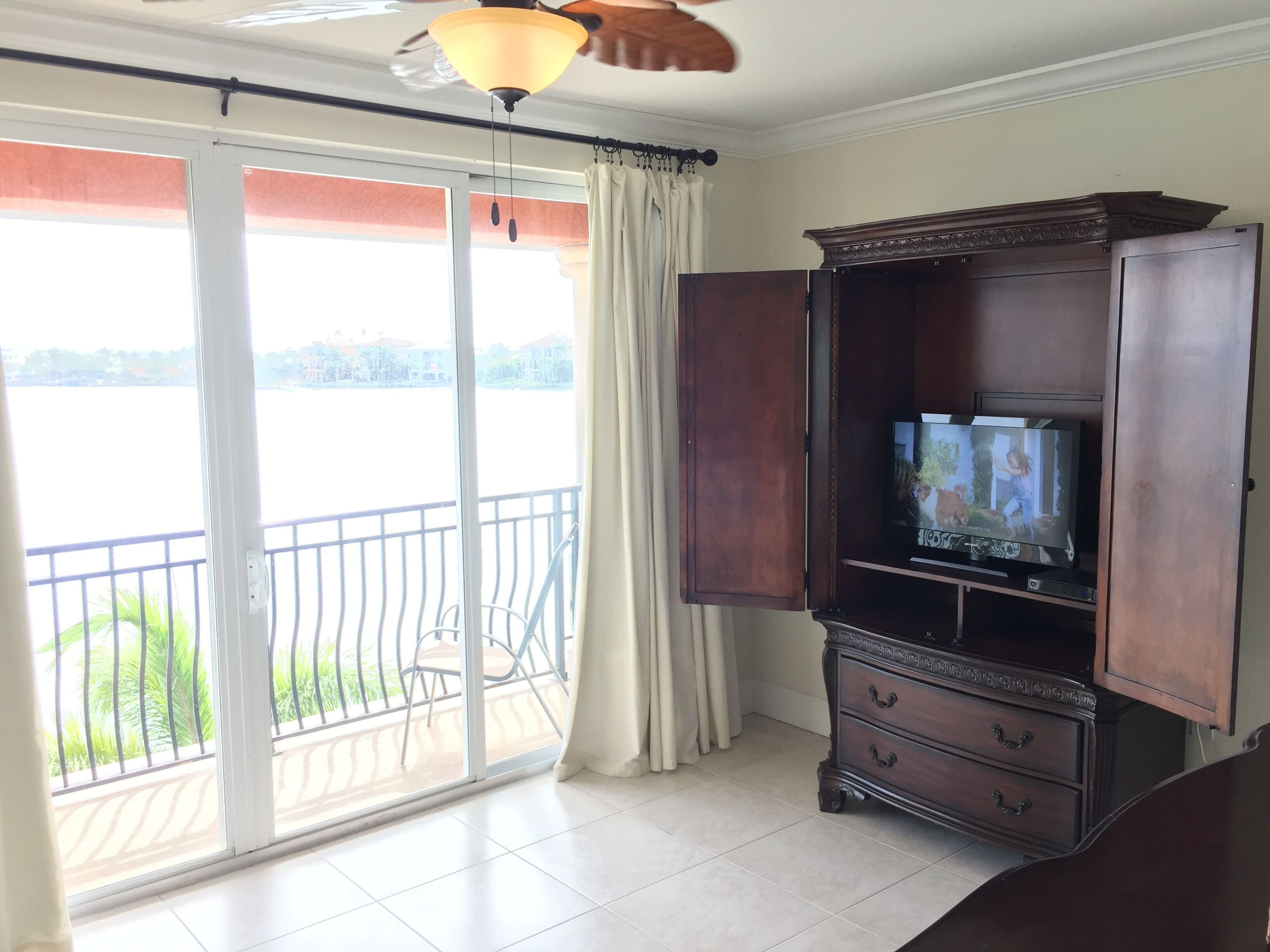 Una habitación con un gran ventanal corredero que da a un balcón, un mueble de madera con un televisor y un ventilador de techo.