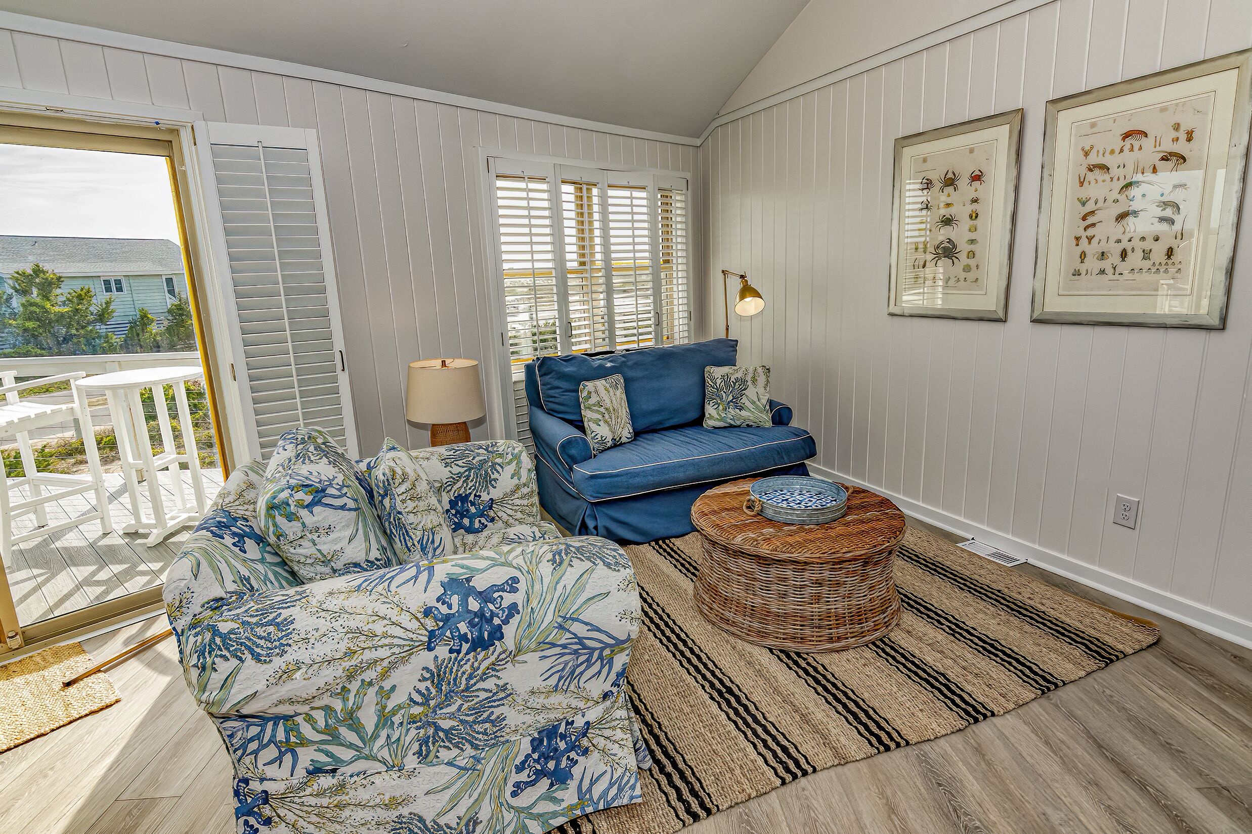 Una sala de estar con un sofá azul, un sillón floral, una mesa de centro de mimbre y un amplio ventanal con contraventanas.