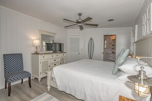 Een slaapkamer met een bed, een stoel, een dressoir, een plafondventilator en een nachtkastje met een lamp.