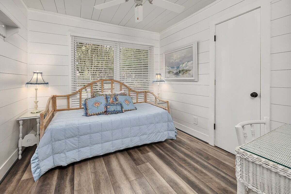 A bedroom with a bed, two bedside lamps, a window with blinds, a door, and a wicker table.