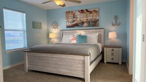 Um quarto com cama de madeira, duas mesas de cabeceira com luminárias, uma janela com persianas, um volante de navio decorativo na parede e uma pintura de uma paisagem portuária.
