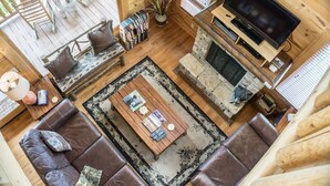 A cozy living room with a stone fireplace, leather sofas, a coffee table with magazines, and a TV.