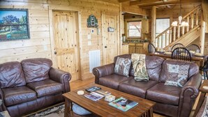 A cozy cabin interior with two leather sofas, a wooden coffee table, and a staircase leading to a kitchen area.