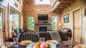 A cozy wooden cabin interior with a dining table set for two, a fireplace, and a television mounted above it.