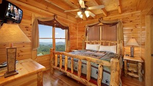 A wooden cabin room with a bed, a ceiling fan, a television, and a view of the mountains.