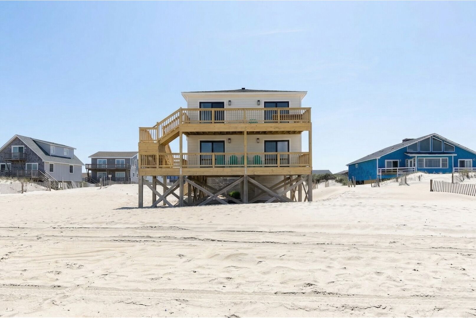 A multi-story wooden structure with a balcony, situated on a sandy beach with other buildings in the background.