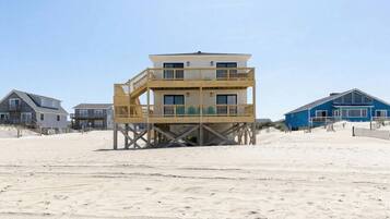 A multi-story wooden structure with a balcony, situated on a sandy beach with other buildings in the background.