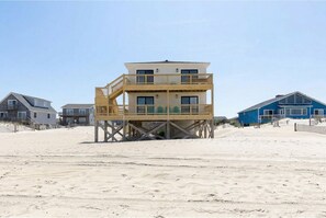 A multi-story wooden structure with a balcony, situated on a sandy beach with other buildings in the background.