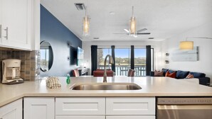 A modern kitchen with white cabinets, a stainless steel sink, and a built-in dishwasher. There is a dining area with a table and chairs, and a large window offering a view of the outdoors. - 2 STORY WITH 2 PRIVATE BALCONIES! 150 STEPS FROM BEACH! (Miramar Beach)