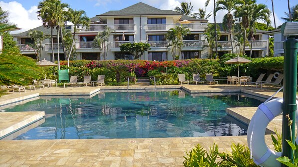 A pool area with lounge chairs, a no swimming sign, and a building with balconies and a garden in the background.