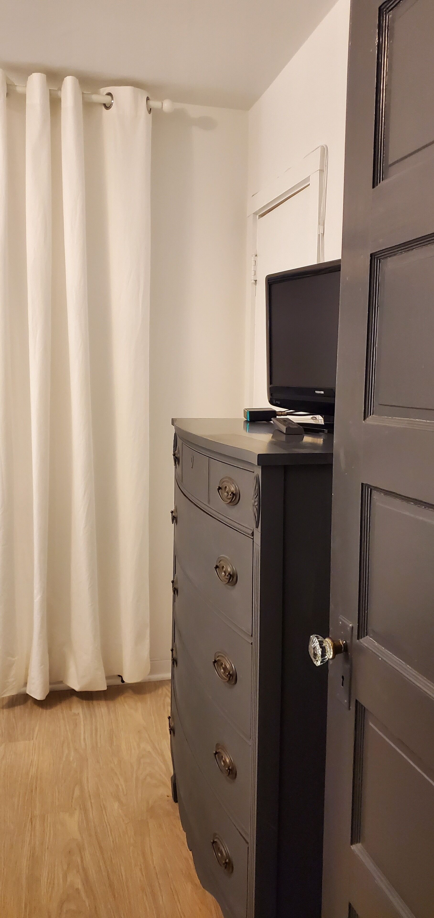 Habitació amb terra de fusta, un taulell negre amb calaixos, una televisió a la part superior i una porta amb ull de cerradura.