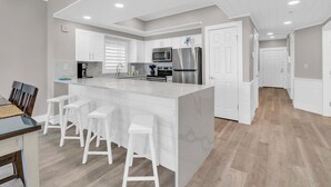Una cocina moderna con armarios blancos, una isla central con taburetes de bar, electrodomésticos de acero inoxidable e iluminación empotrada en el techo.